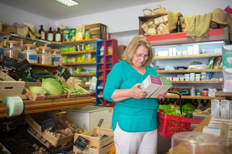 A shopper in a grocery store aisle holds a packaged food item and reads its label.